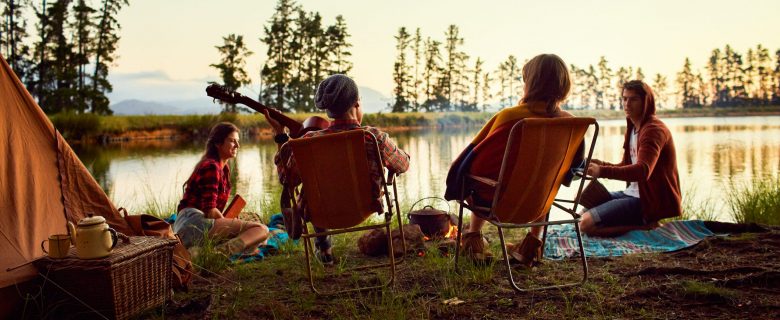 Four friends, one playing guitar, enjoy the sunset at their lakeside campground.