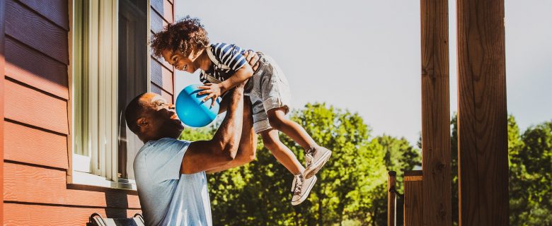 A smiling father holds up his son who has a blue ball in his hands. 