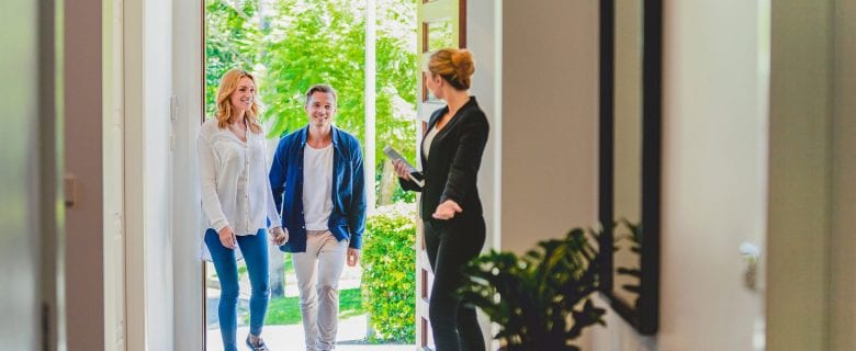 A realtor stands at the front door of a house, welcoming a couple inside.