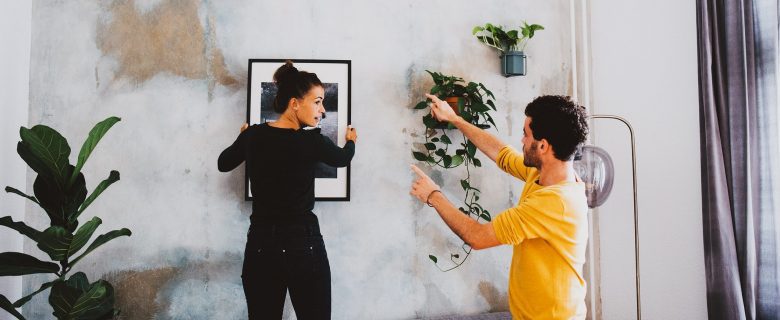 A woman holds a picture frame up to the wall and turns to face a man who is offering her tips on placement. 