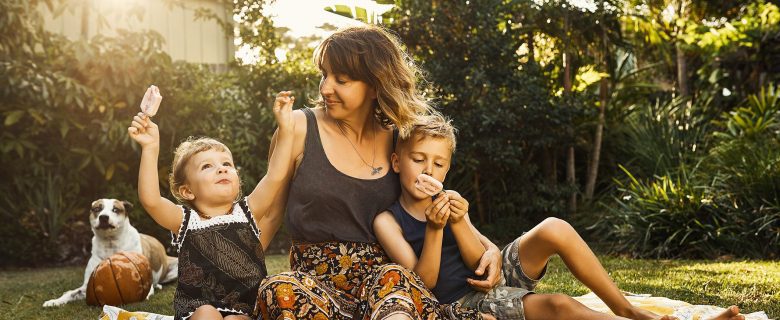 A mother sits on a blanket with two kids holding popsicles. A dog is sitting behind them on the grass. 