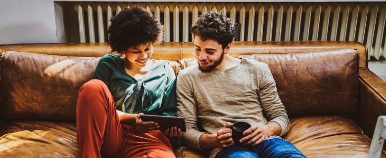 A man and woman sit together on a brown leather couch looking at a tablet together.