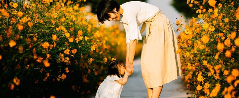 A woman bends down and holds the hands of a toddler in the middle of a path that is lined on either side with flowers.
