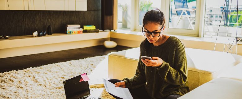 A woman sits on the floor next to a sectional couch looking at her phone and holding a notebook. 