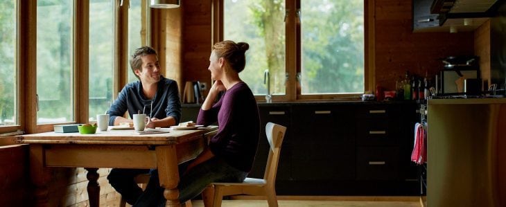 A man and woman talk to teach each other at a kitchen table. There are mugs and plates of cake on the table. 