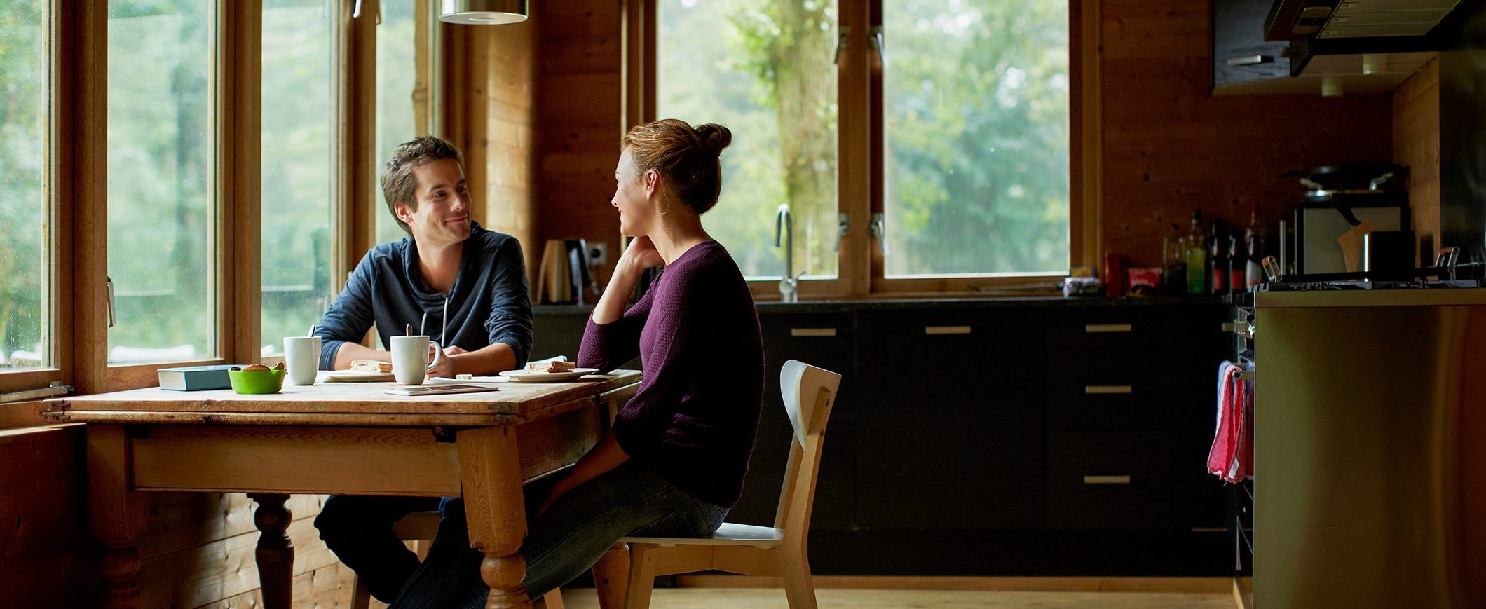 A man and woman talk to teach each other at a kitchen table. There are mugs and plates of cake on the table.