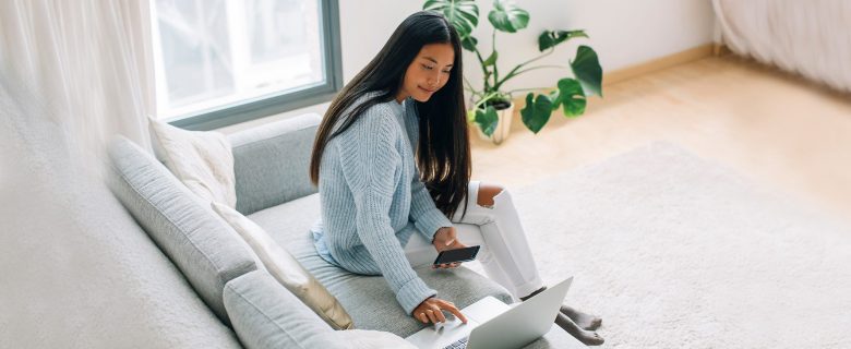 A woman sitting on her couch, holding a smart phone and using a laptop computer.