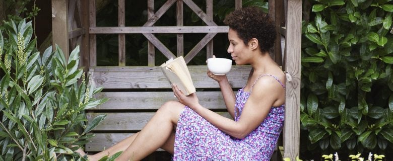 A woman sits in a garden, reading a book and sipping a cup of coffee.