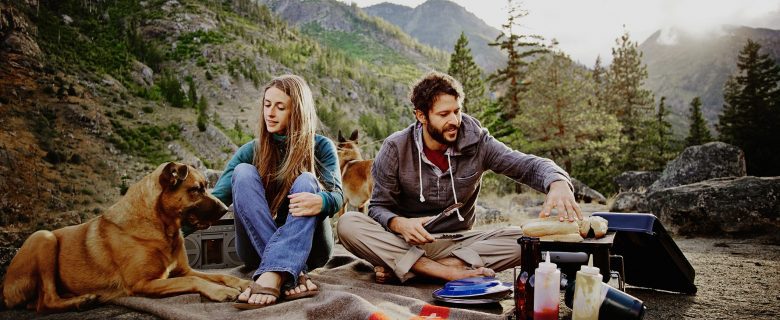 A couple, camping with their dogs, sets up camp and cooks a meal.