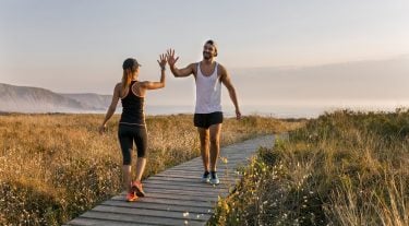 A man and a woman in workout gear high five in a field by the ocean.