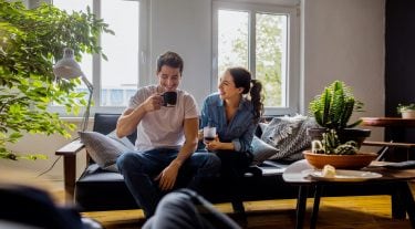 A man and a woman sit on a couch in their sunlit apartment. They are drinking coffee.