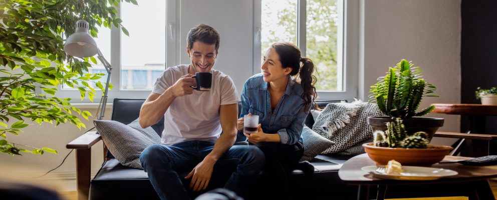 A man and a woman sit on a couch in their sunlit apartment. They are drinking coffee.