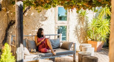 A woman wearing a red romper sits outside on a couch and looks at a tablet.