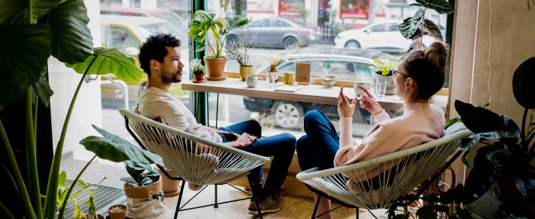 A man and woman sit at a table in front of a window talking.