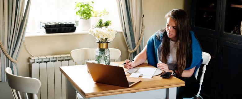 A woman sits at a table facing a laptop and reviewing paperwork, pen in hand.