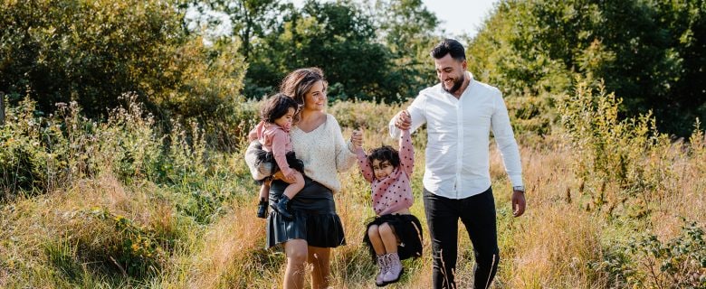 A mother and father walk through a field. The mother carries a small child, and both parents hold the hands of a second child.