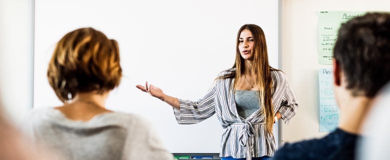 A woman speaking while standing in front of a white board, with two people listening.