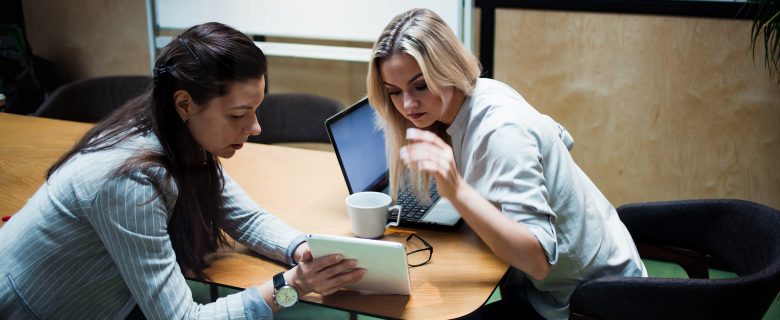 A woman showing a co-worker something on a tablet, while the co-worker sits in front of a laptop and a coffee mug.