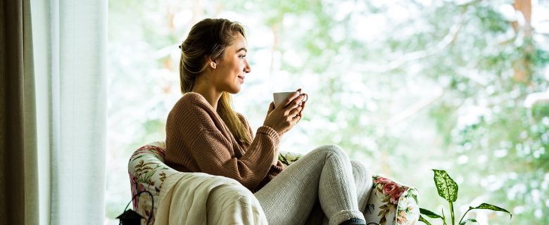 A woman sitting on a chair with a cup of coffee, looking out the window.