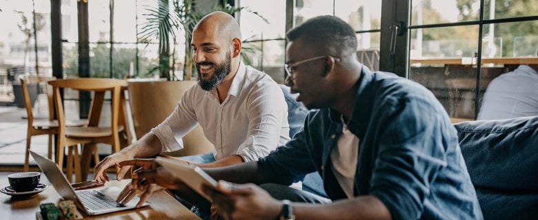 Two men sit at a table looking at a laptop.