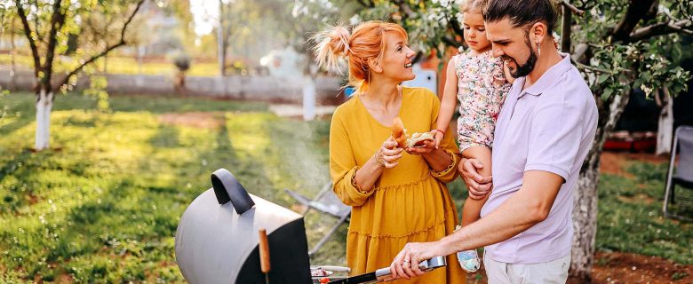 A man holds his daughter while grilling as the mother stands nearby and smiles.