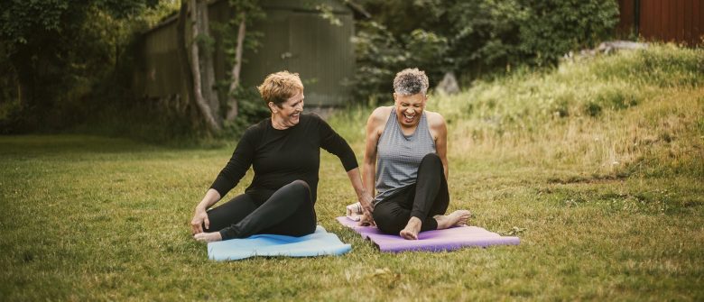 Two laughing women sit on yoga mats outside on the grass.