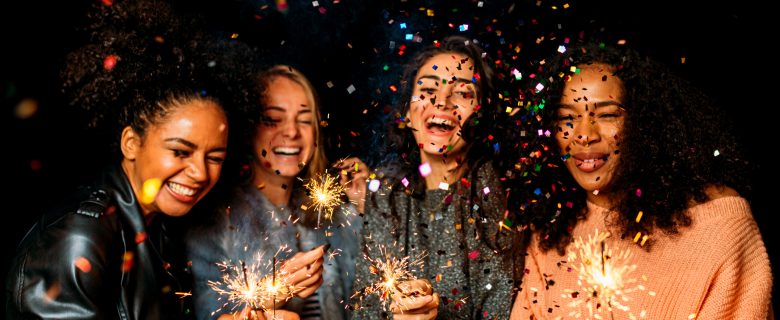 Four women laugh and hold sprinklers as confetti falls around them.