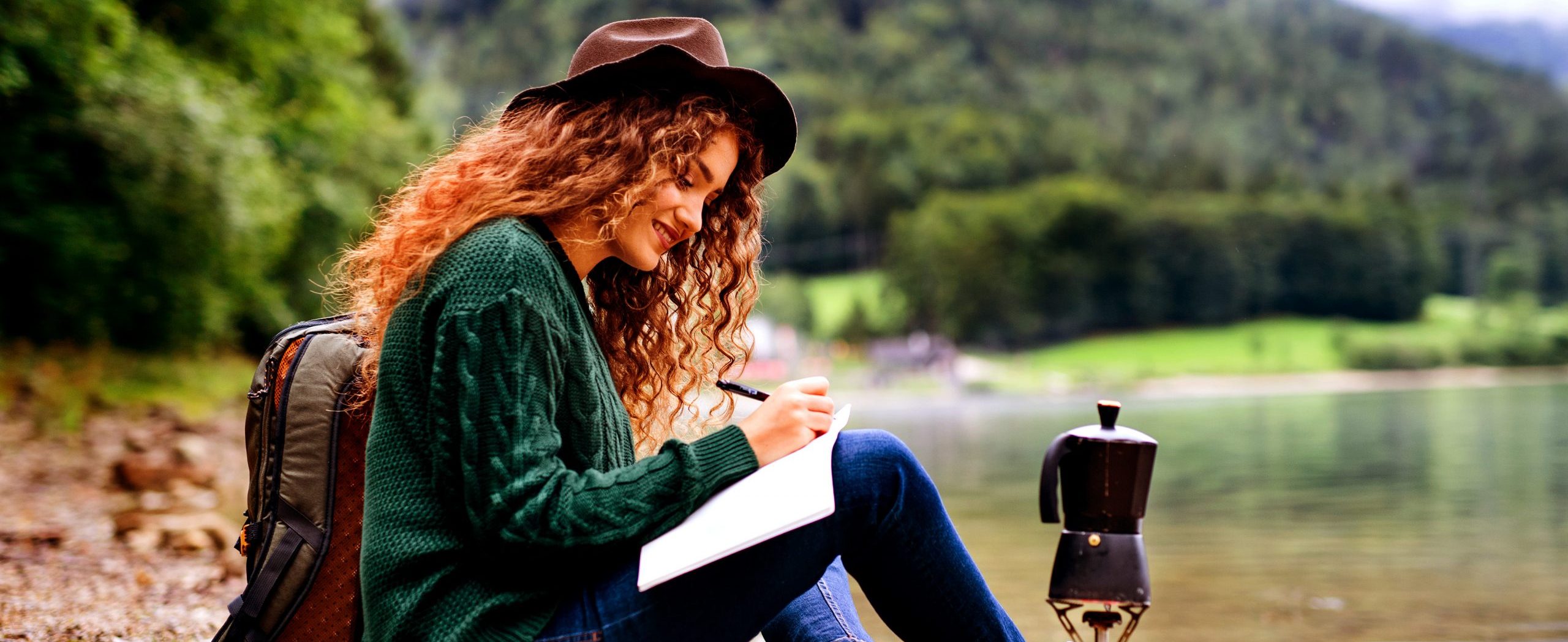 A woman sits by the edge of a lake writing on a piece of paper.