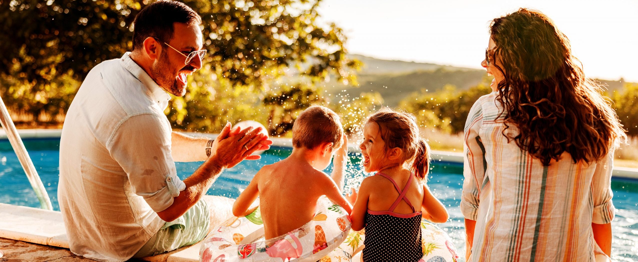 A dad and mom sit at the edge of a pool with their young son and daughter.