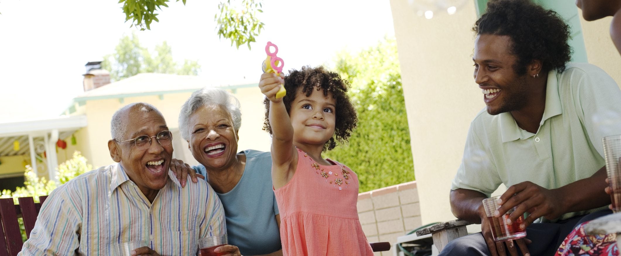 A girl plays with a bubble gun as her father and grandparents look on smiling.