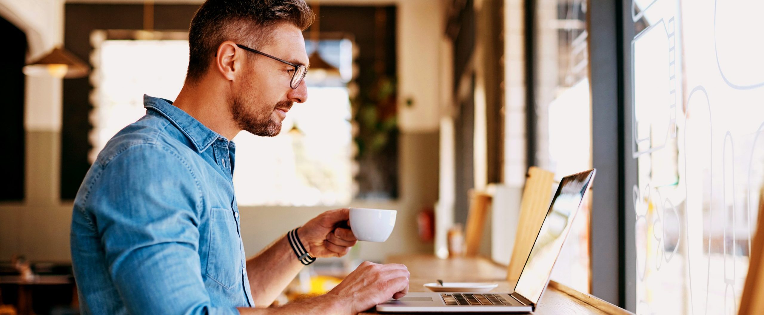 A man drinks a cup of coffee and looks at his laptop.