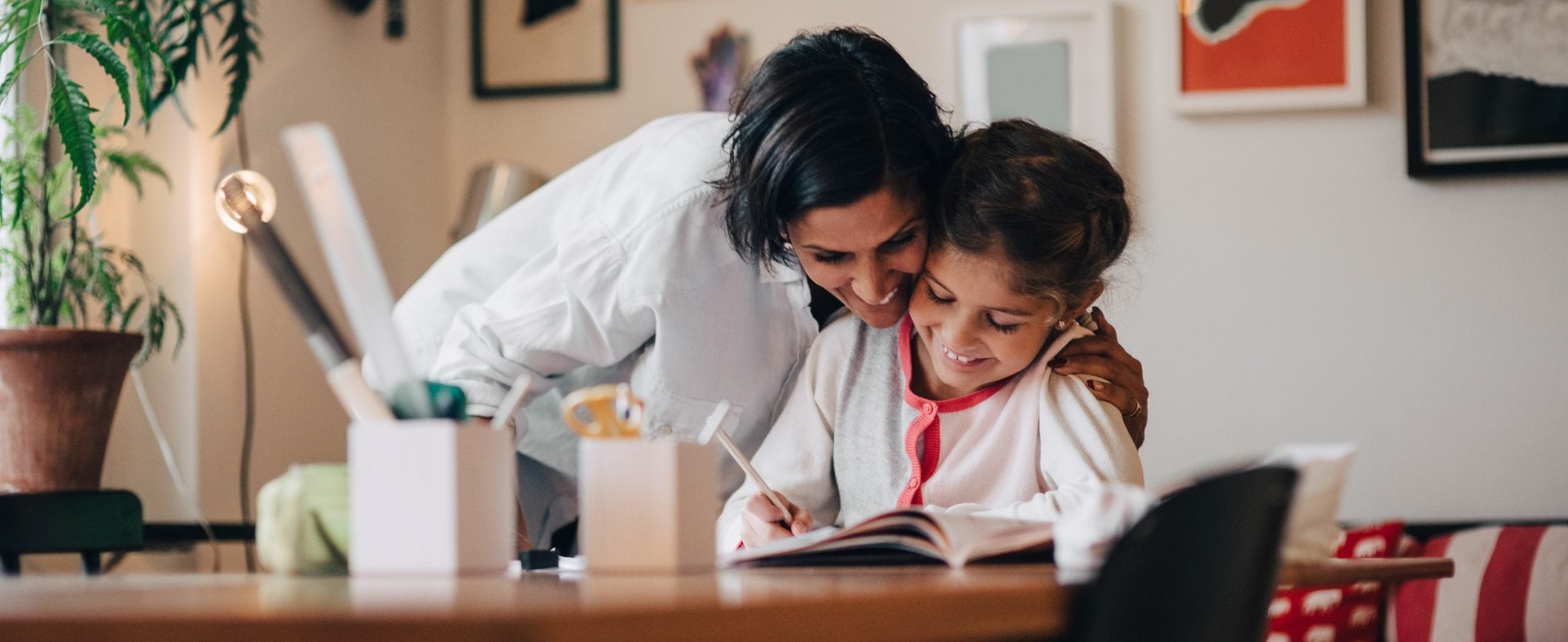 A girl writing in a notebook while being hugged by her mother.