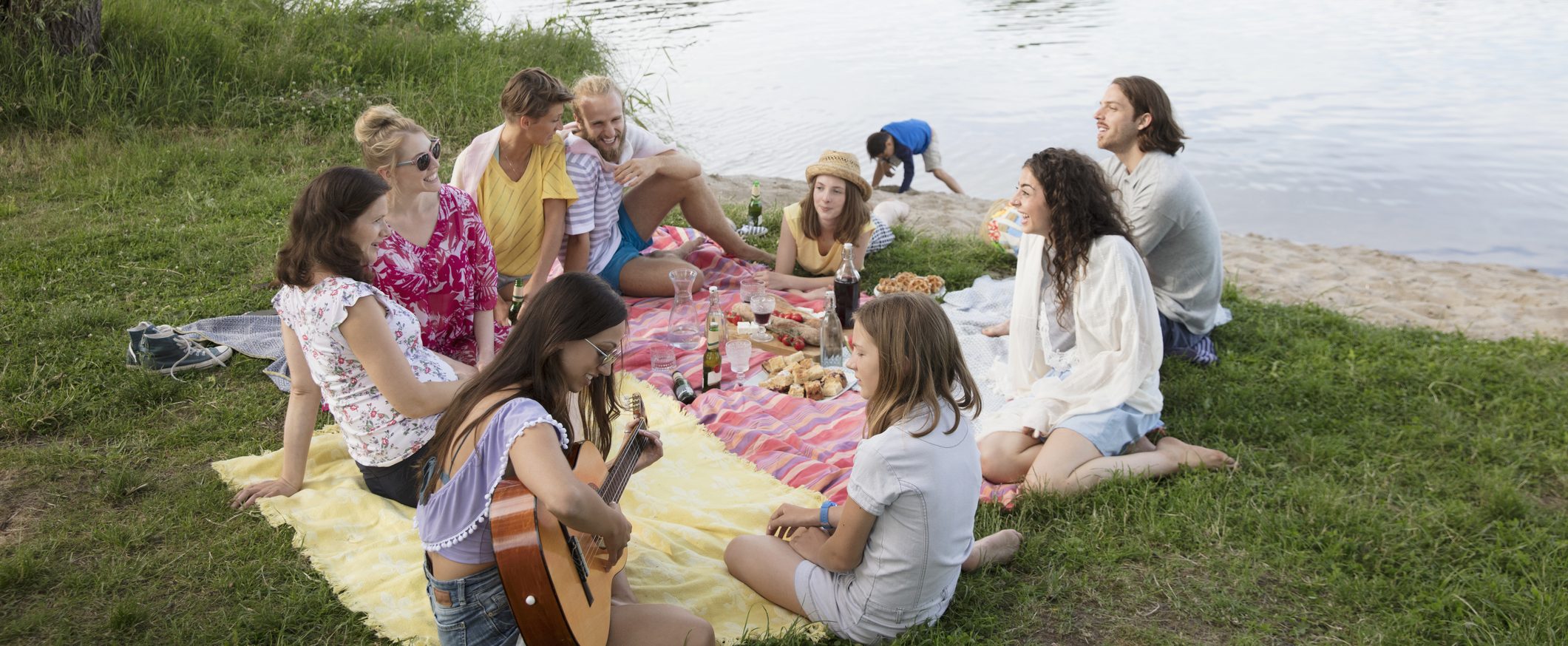 A group of 10 young people enjoying a picnic on grass, near the edge of a lake.