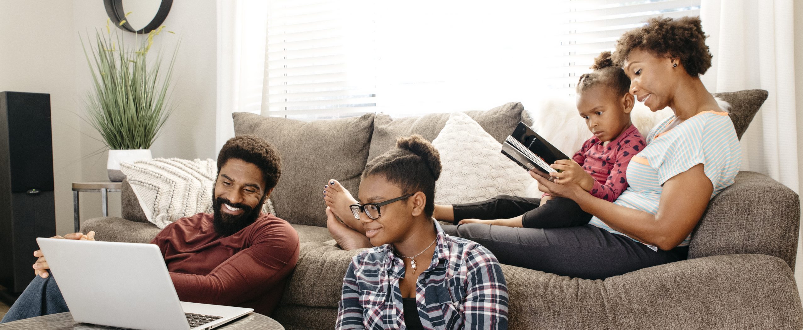 Two individuals sitting on the floor looking at a laptop screen, while a mother reads a book to her daughter sitting on a sofa in the background.