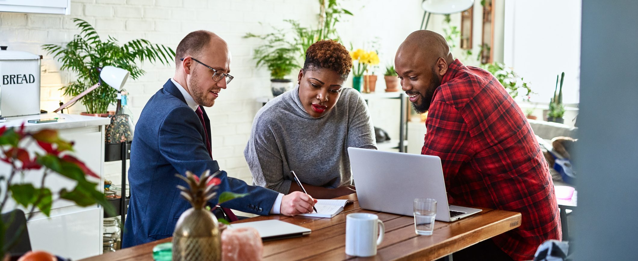 A financial advisor speaks to a couple, writing something in a notepad. There is an open laptop on the table in front of them.