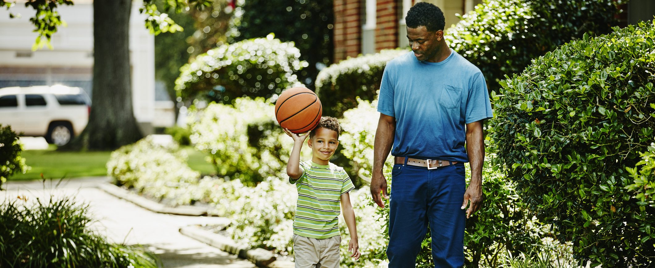 Man proudly looks at his son playing with a basketball as they walk through the neighborhood