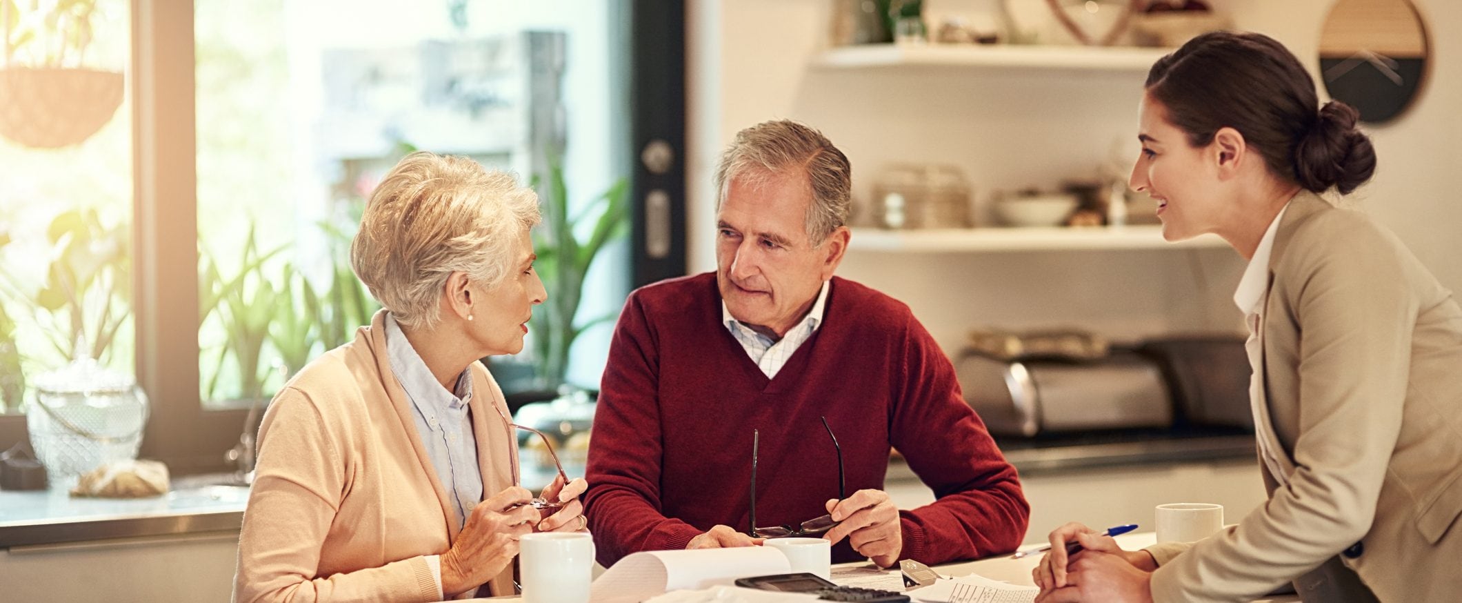 An elderly couple sits at a kitchen island that is covered with papers and coffee mugs. A woman in a suit jacket looks at them, smiling.