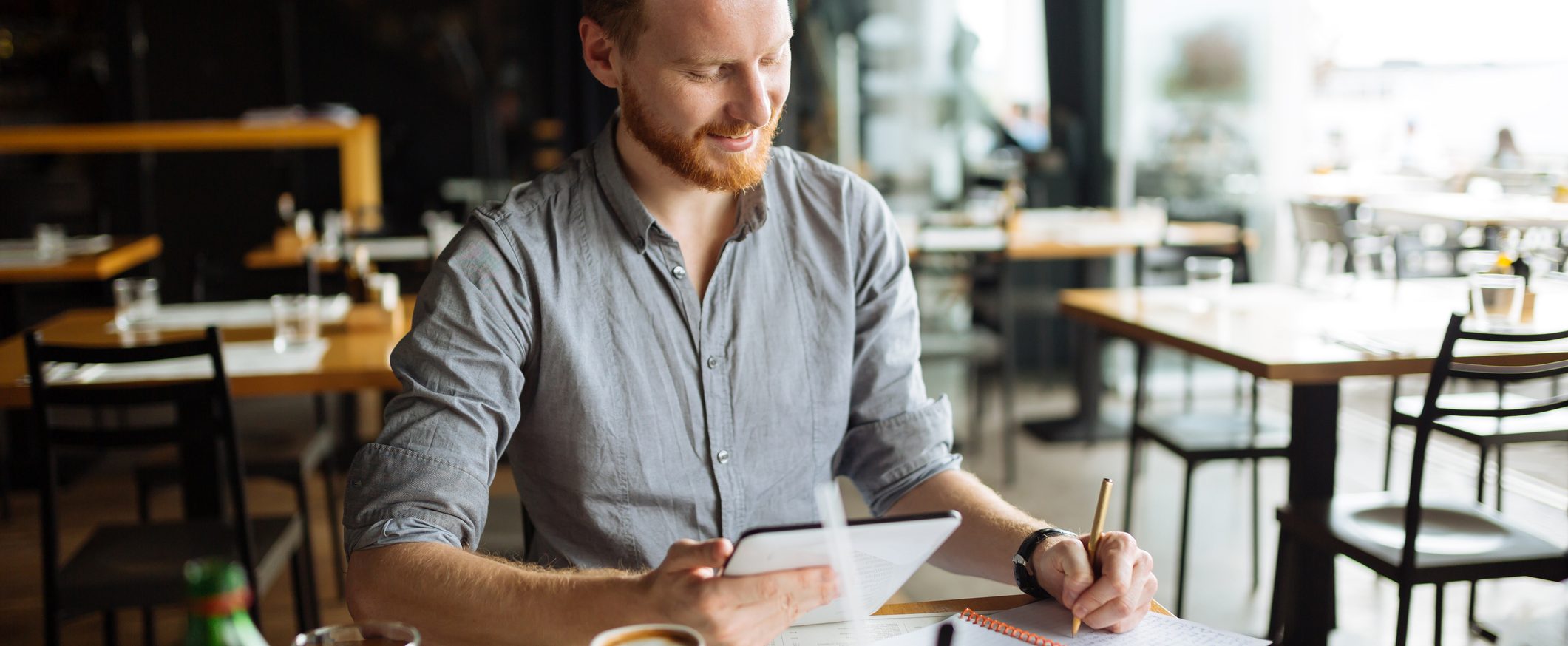 A man sits at a table in a cafe, holding a tablet in one hand and writing in a notebook with the other hand.