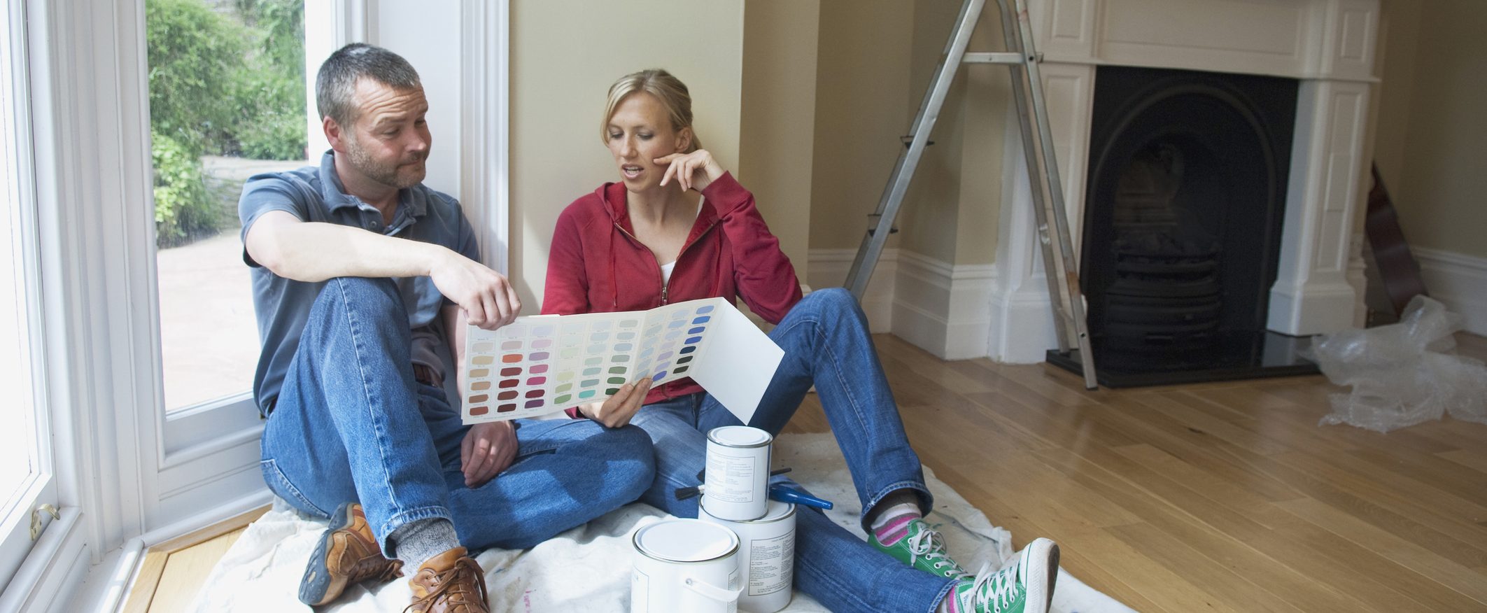 An image of a couple sitting on the floor of a room under renovations looking at brochures of different paint colors.