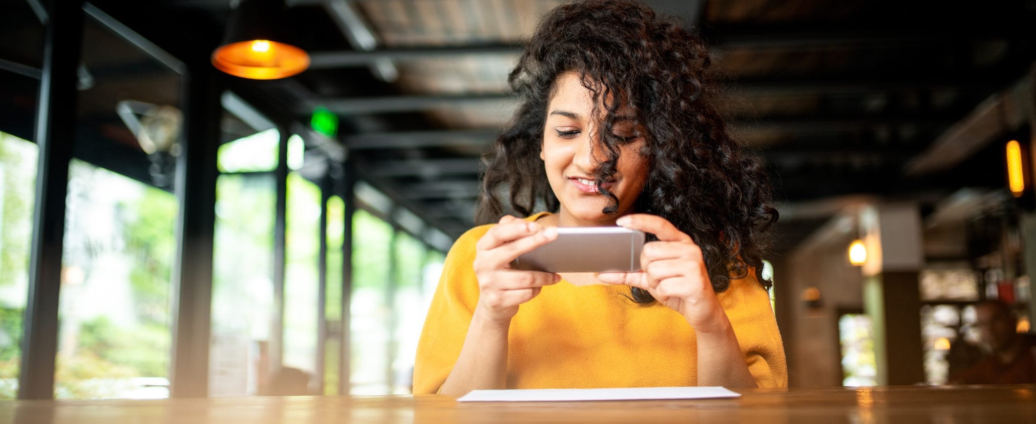 A woman sits at a table and takes a picture of a check with her smart phone.