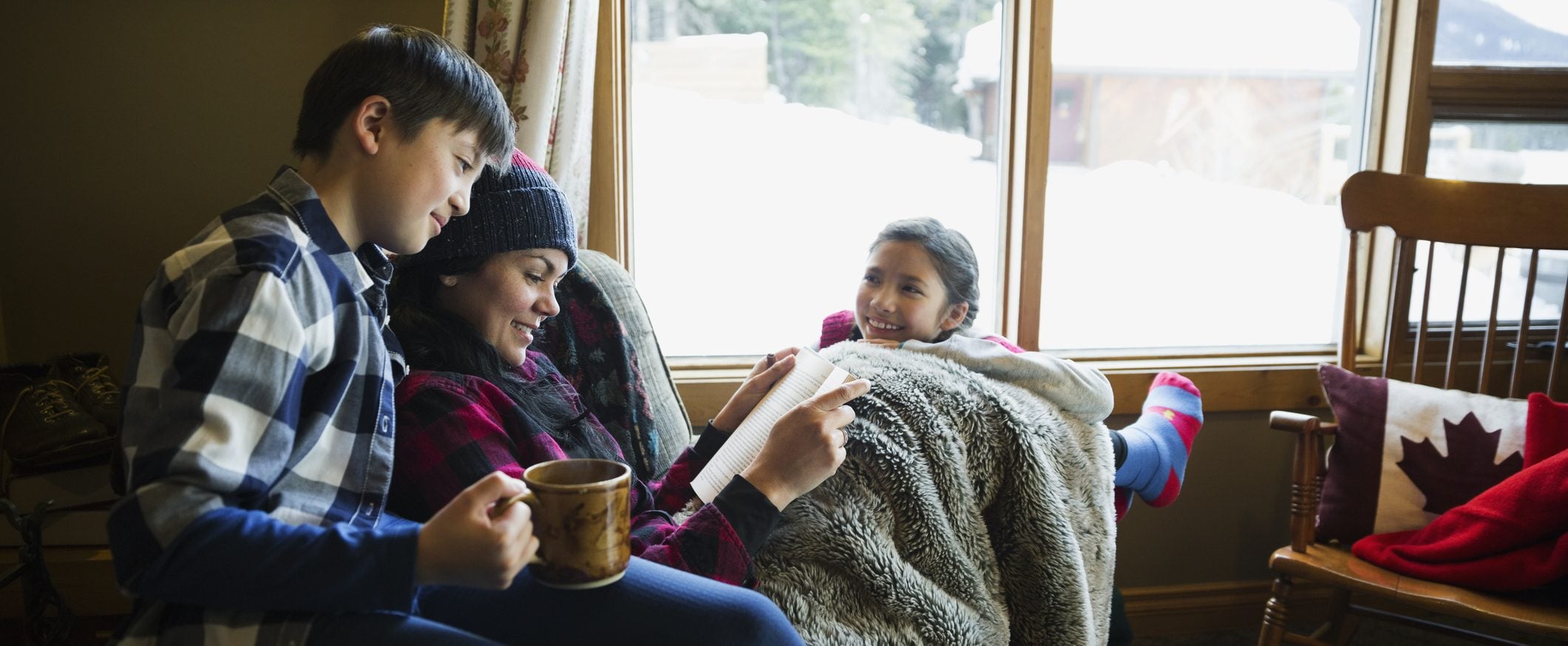 A woman sits near a window that looks out on a snowy landscape. She holds a book, and a child sits on either side of her.