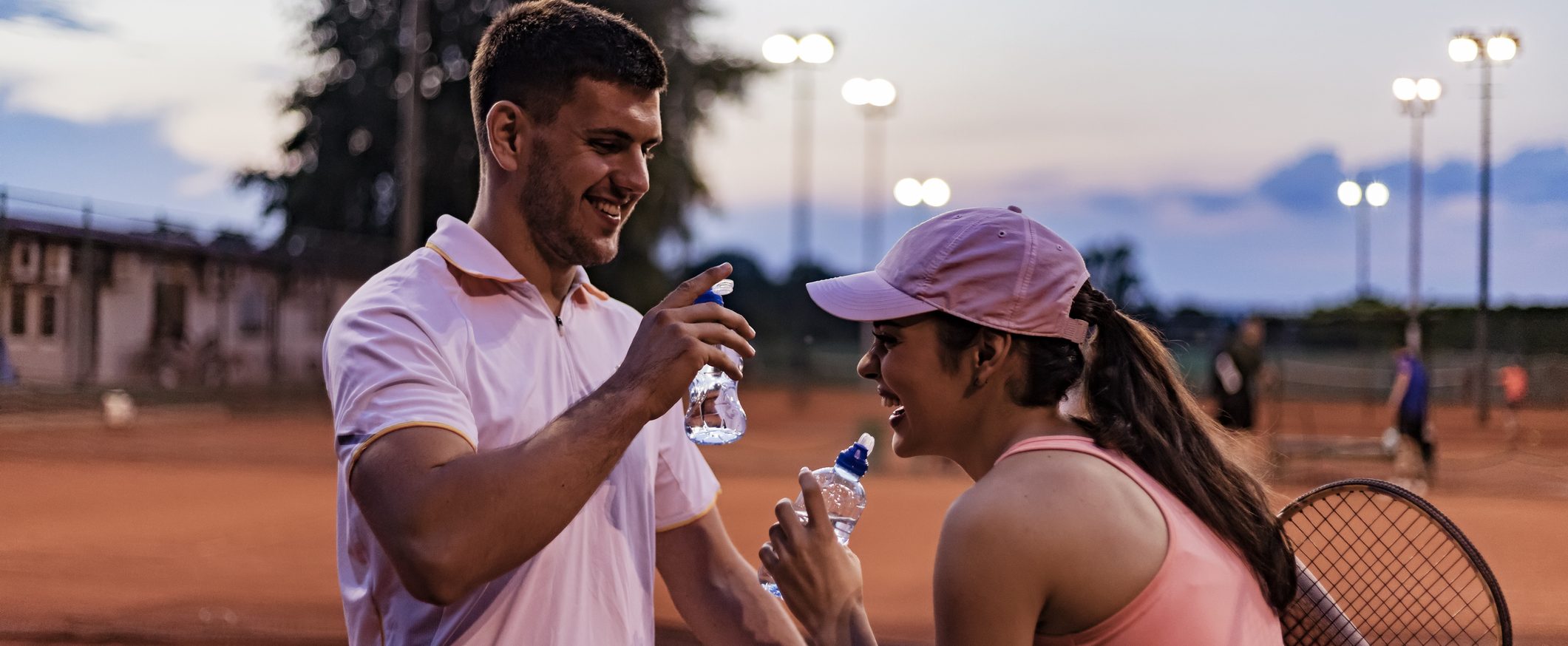 A couple takes a break from playing tennis. They smile and hold water bottles.