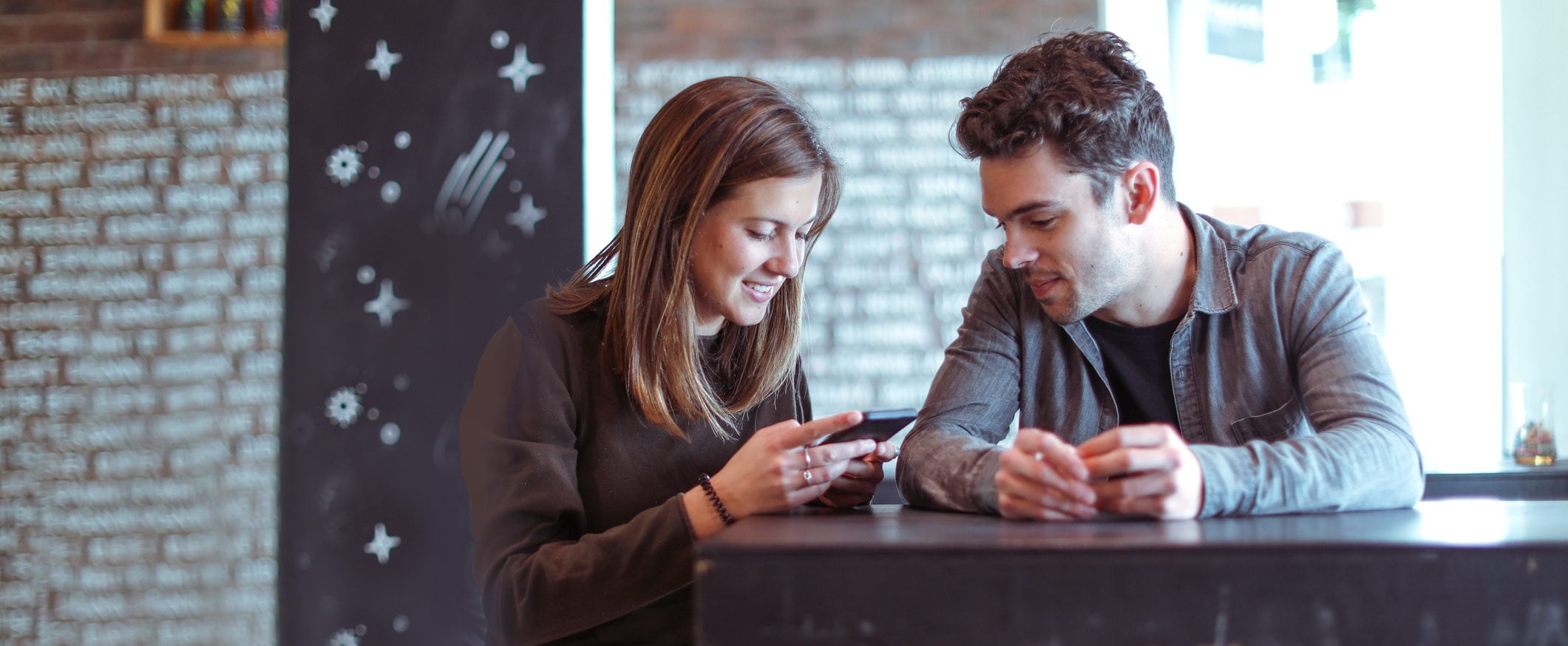 A couple sits at a counter together, smiling at the screen of a smart phone.