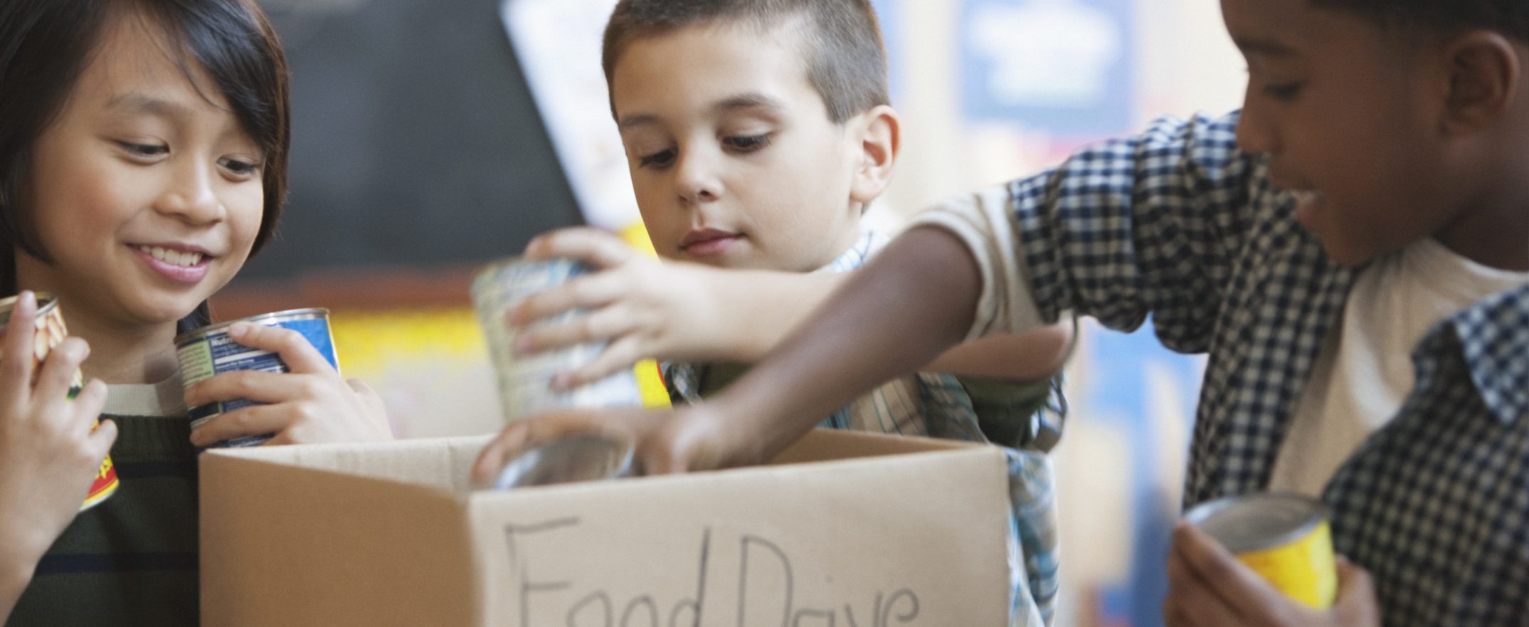 Three young kids place cans of food into a cardboard box marked “Food Drive.”