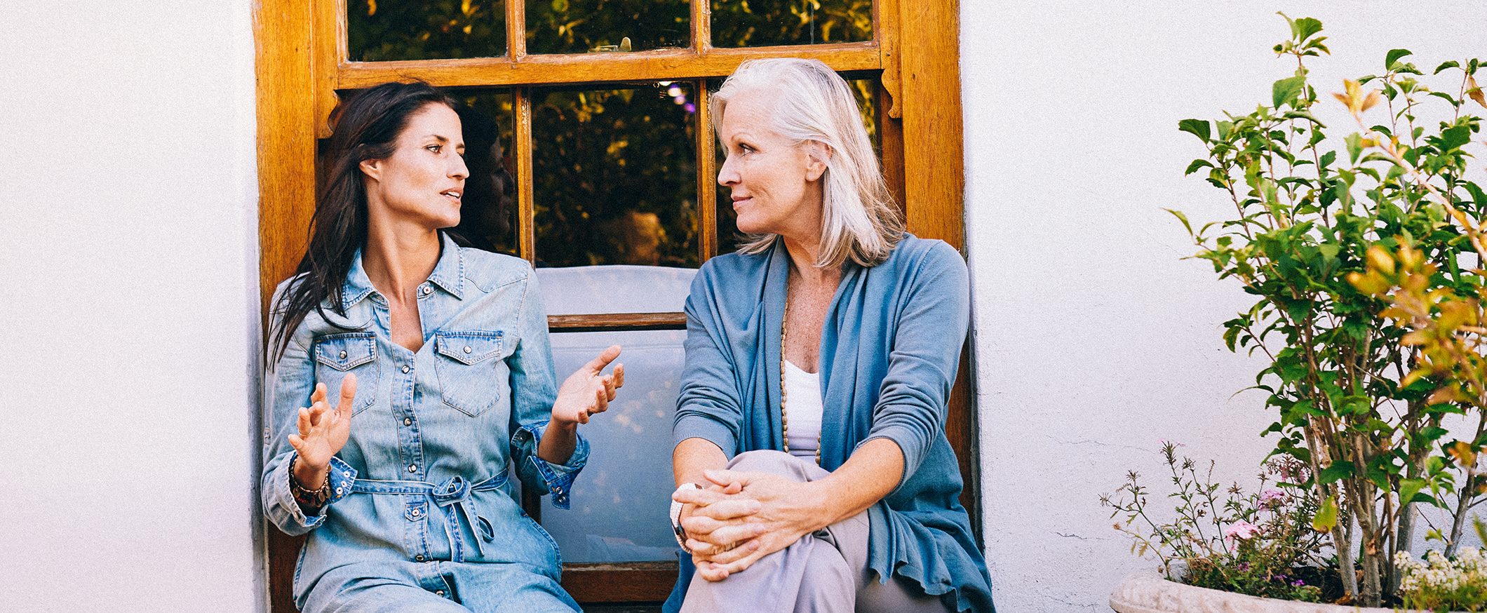 A younger woman with dark hair sits outside in front of a window with an older woman with graying hair. There are potted plants next to them.