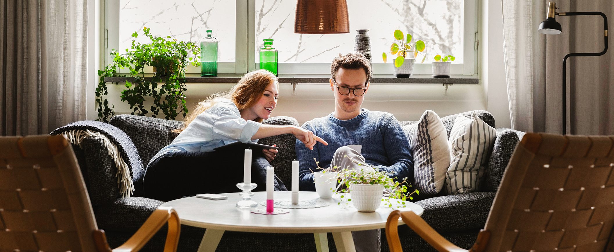 A man and a woman sit together on a couch in front of a window, looking at something on a phone. There are plants and green bottles and a vase on the windowsill.