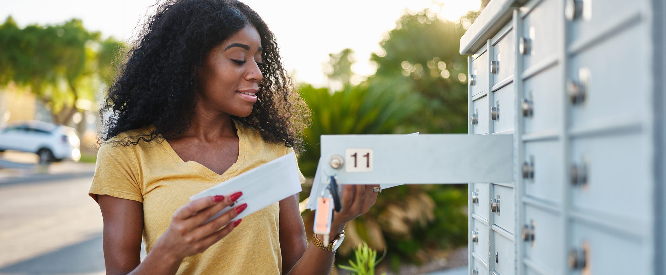 A woman wearing a yellow t-shirt opens her mailbox with a key and looks at some mail.