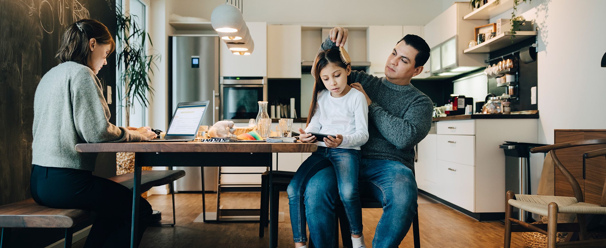 A family sits at a kitchen table in a home. The mom works on a laptop, while a daughter sits on her dad’s lap while he brushes her hair.