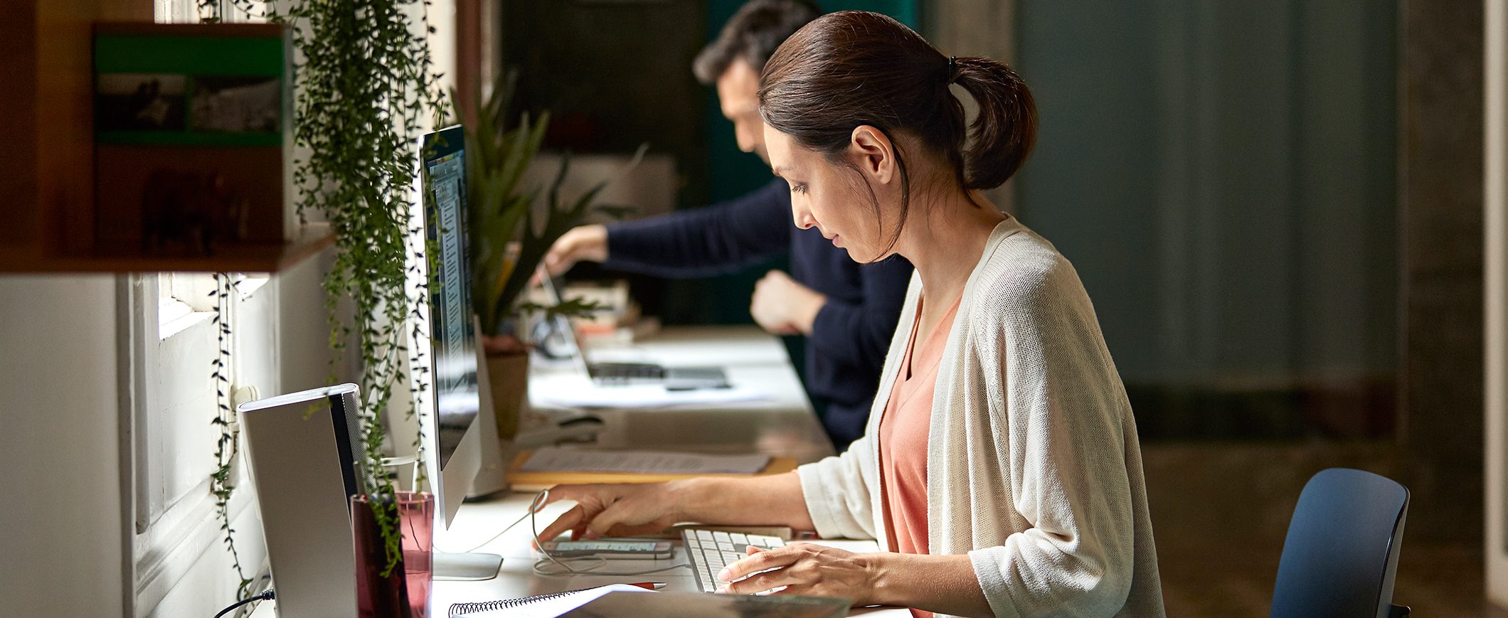 A woman sits at a home office looking at a desktop computer and phone. A man shares the same desk, working on a laptop.