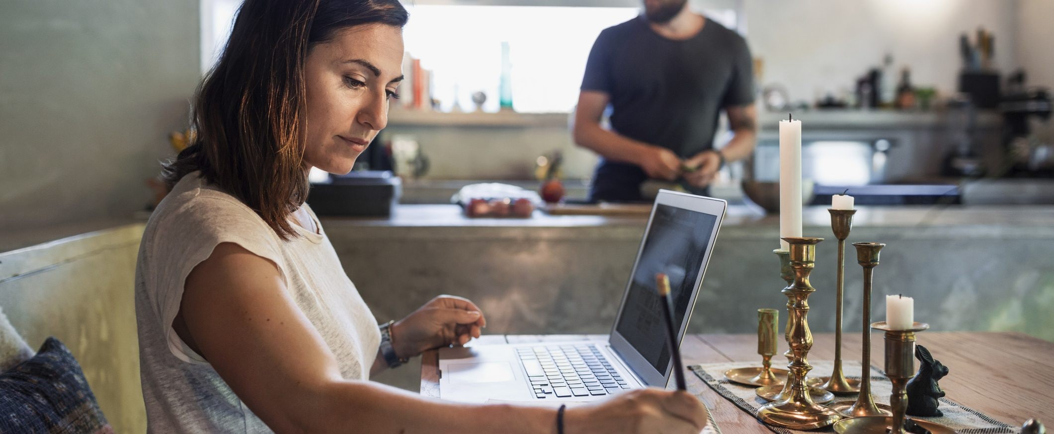 A woman sits at a table in a kitchen, working on a laptop and writing something with a pencil. A man in the background prepares food at a counter.